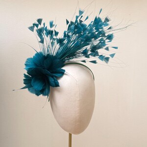 May include: A teal fascinator headpiece featuring a dense cluster of soft, layered feathers at the base, accented by long, thin stems topped with small, delicate feather tufts that extend outward in a fan-like arrangement on a thin headband.