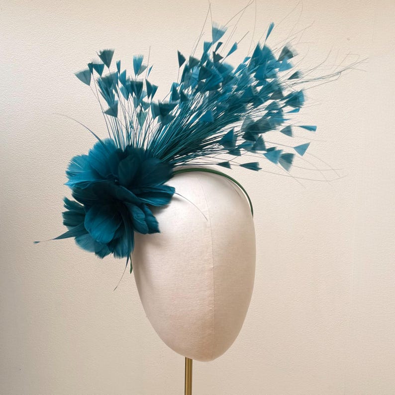 May include: A teal fascinator headpiece featuring a dense cluster of soft, layered feathers at the base, accented by long, thin stems topped with small, delicate feather tufts that extend outward in a fan-like arrangement on a thin headband.