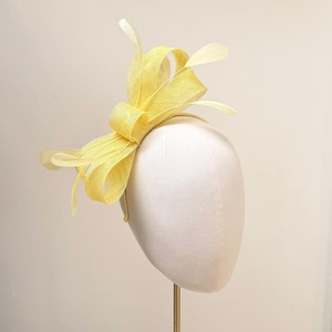 May include: A bright yellow fascinator headpiece featuring a structured bow made of stiffened mesh fabric. The design includes several delicate, thin feathers extending outward from the center, all mounted on a thin, matching yellow headband.