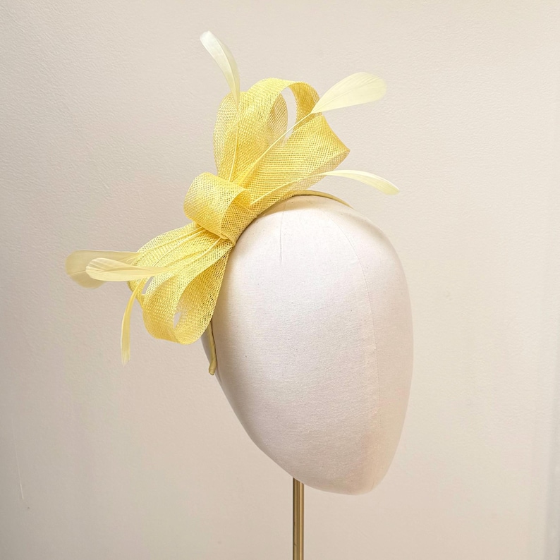 May include: A bright yellow fascinator headpiece featuring a structured bow made of stiffened mesh fabric. The design includes several delicate, thin feathers extending outward from the center, all mounted on a thin, matching yellow headband.