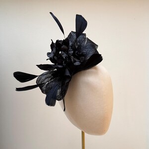 May include: A black fascinator featuring a cluster of fabric flowers, feathers, and netting. The headpiece is mounted on a neutral-toned form, suggesting a formal occasion accessory. The design incorporates various textures and shapes.