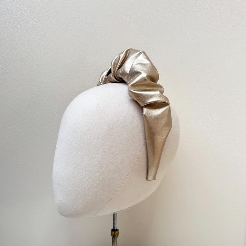 May include: A gold-colored headband made of faux leather with a gathered, knotted design.