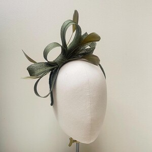 May include: A green headpiece with a bow and feather accents. The headpiece is made of a textured fabric and is attached to a thin headband. The bow is large and has several loops. The feathers are a darker shade of green.