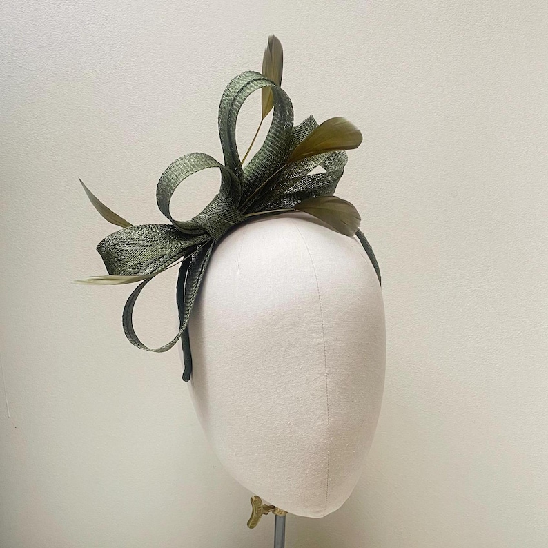 May include: A green headpiece with a bow and feather accents. The headpiece is made of a textured fabric and is attached to a thin headband. The bow is large and has several loops. The feathers are a darker shade of green.