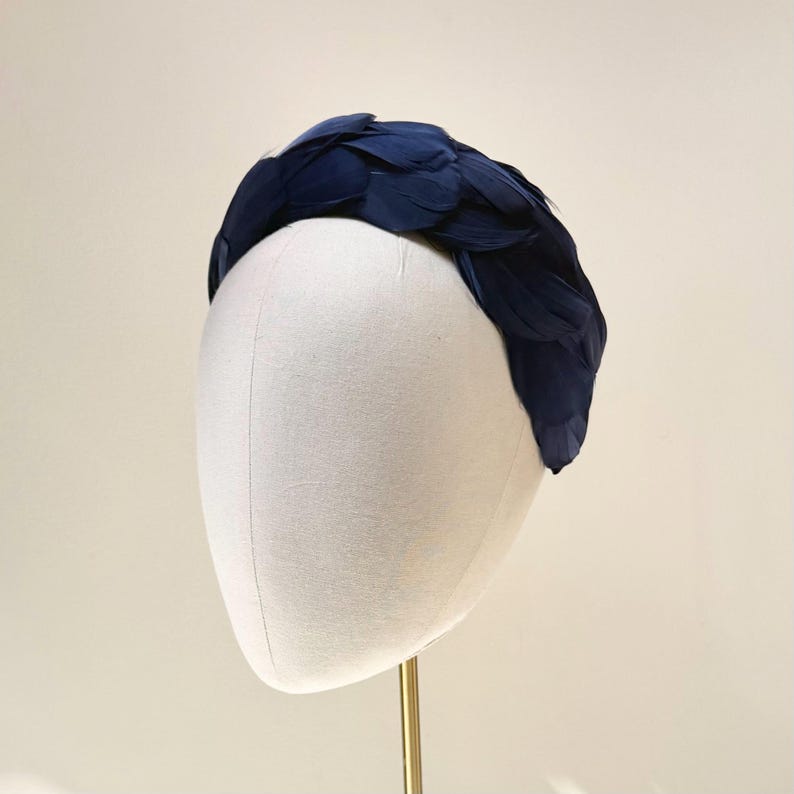May include: A wide headband completely covered in layered, dark navy blue feathers. The feathers are arranged in a dense, overlapping pattern that creates a textured, voluminous appearance across the entire surface of the curved hair accessory.