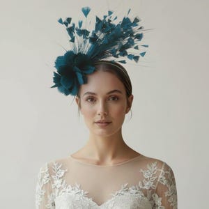 May include: A teal fascinator headpiece featuring a cluster of soft fabric petals and an arrangement of upright, wispy feathers. The headpiece is secured on a thin, matching teal headband, creating a structured and decorative hair accessory.