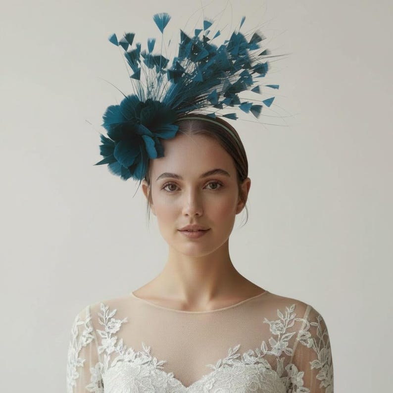 May include: A teal fascinator headpiece featuring a cluster of soft fabric petals and an arrangement of upright, wispy feathers. The headpiece is secured on a thin, matching teal headband, creating a structured and decorative hair accessory.