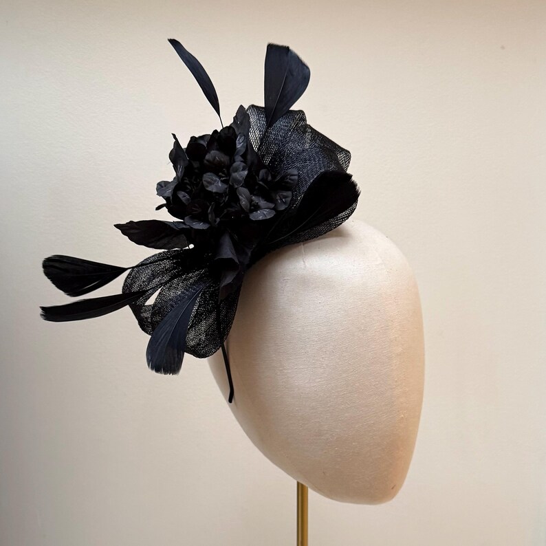 May include: A black fascinator with a floral design and feathers. The headpiece is displayed on a mannequin head. The fascinator features a cluster of fabric flowers and several black feathers. The base is a black mesh.
