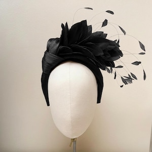 May include: A black headband featuring a wide, knotted fabric band and a large cluster of black feathers on one side. The design includes long, thin, wispy feather accents that extend outward, creating a textured and layered appearance.