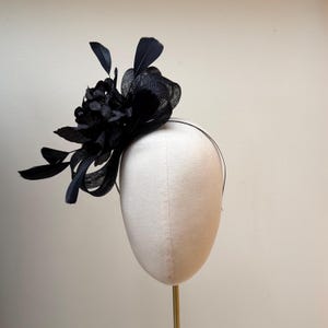 May include: A black fascinator featuring a cluster of fabric flowers, feathers, and mesh detailing. The headpiece is mounted on a thin, black headband. The design is elegant and suitable for formal events.