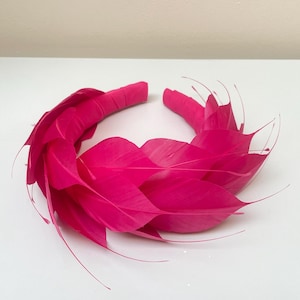 May include: A bright pink feather headband. The headband is made of a thick band of fabric covered in feathers. The feathers are arranged in a circular pattern around the headband.