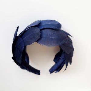 May include: A navy blue headband adorned with a dense arrangement of layered, smooth feathers. The feathers are arranged in an overlapping pattern, creating a textured, circular crown shape that covers the entire band from end to end.