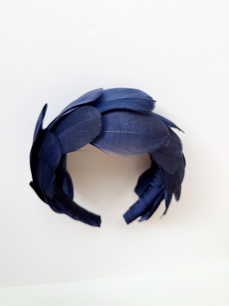 May include: A navy blue headband adorned with a dense arrangement of layered, smooth feathers. The feathers are arranged in an overlapping pattern, creating a textured, circular crown shape that covers the entire band from end to end.