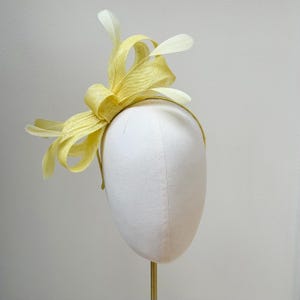 May include: A pale yellow fascinator headpiece featuring a structured bow made of woven material, accented with several delicate, matching pale yellow feathers extending outward from the center, all mounted on a thin, circular headband frame.
