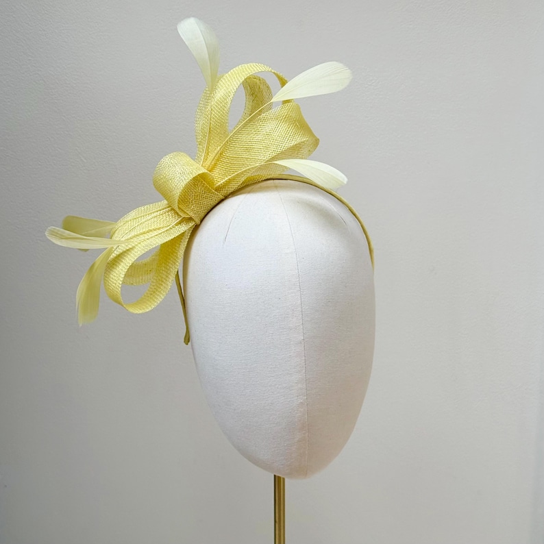 May include: A pale yellow fascinator headpiece featuring a structured bow made of woven material, accented with several delicate, matching pale yellow feathers extending outward from the center, all mounted on a thin, circular headband frame.