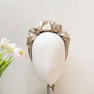May include: A gold-colored headband made of faux leather with a gathered, ruffled design. The headband is displayed on a white mannequin head.