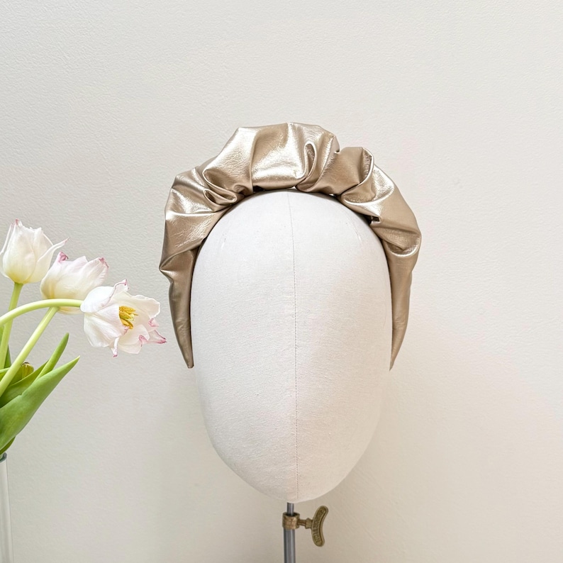 May include: A gold-colored headband made of faux leather with a gathered, ruffled design. The headband is displayed on a white mannequin head.