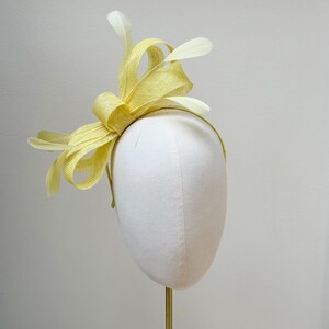 May include: A pale yellow fascinator headpiece featuring a structured bow made of woven material, accented with several delicate, matching pale yellow feathers extending outward from the center, all mounted on a thin, circular headband frame.