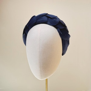May include: A dark navy blue headband covered in layered, overlapping feathers. The accessory features a rounded, structured shape that sits atop a neutral-colored display form, showcasing a dense arrangement of individual dark blue plumage.