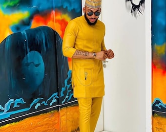 May include: A mustard yellow two-piece suit with a long-sleeved tunic and matching trousers. The outfit includes a small pocket on the tunic and is paired with black loafers. The person is wearing a patterned cap and sunglasses.