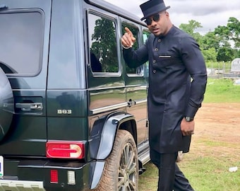 May include: A black Mercedes-Benz G-Class SUV with the B63 model designation. The vehicle is parked on grass. A person wearing a black outfit, sunglasses, and a top hat is standing next to the car.