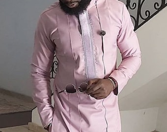 May include: A light pink two-piece suit with a long-sleeved tunic and matching pants. The tunic features vertical pleats, a decorative center panel, and a small brooch. The person is holding sunglasses.