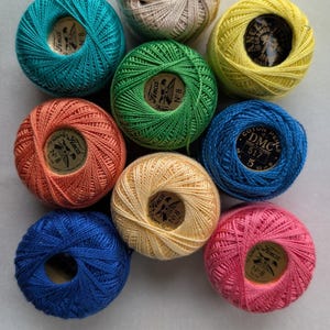 May include: Nine round balls of embroidery thread in various colors including teal, beige, yellow, green, orange, light yellow, blue, dark blue, and pink. Each ball features a central circular label with brand text like Finca and DMC.