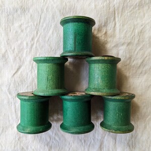 May include: Six vintage wooden spools painted in a vibrant green hue, arranged in a pyramid shape. The spools have a classic cylindrical design with a slightly flared top and bottom. The wood grain is visible, adding to their rustic charm.