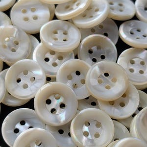 May include: A collection of off-white, pearlescent buttons with four holes each. The buttons are round and have a slightly iridescent sheen, suggesting a mother-of-pearl material. The buttons are scattered, creating a textured surface.