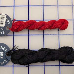 May include: Two twisted skeins of yarn, one bright red and one black, each attached to a circular dark blue tag. The tags feature the white text PLANET EARTH FIBER and SILK PLY, with a small barcode sticker on the red yarn's tag.