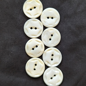 May include: Seven off-white, round buttons with a starburst design and two holes each. The buttons are arranged vertically against a dark background. These buttons are suitable for sewing and craft projects.