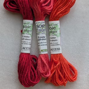May include: Three skeins of Soie d'Alger pure silk embroidery thread in shades of deep red, medium pink, and bright orange. Each 5-meter pull-skein features a white paper label with black text detailing the brand and product specifications.
