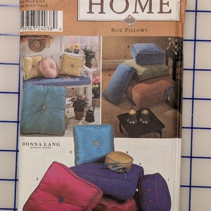 May include: Simplicity Home sewing pattern for box pillows. The pattern cover features images of various colorful box pillows in shades of blue, pink, purple, and gold. The text "Box Pillows" is visible.