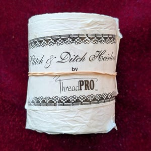 May include: A roll of white fabric with the text "Pitch & Ditch Heirloom by ThreadPRO" printed on the label. The label has a black and white decorative border and is secured with a rubber band. The background is a deep red.