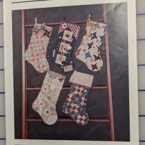 May include: A pattern booklet titled Memory Stockings by thimbleberries featuring five decorative fabric stockings with various quilt-style designs, including star patterns, lace accents, and patriotic motifs, displayed hanging on a wooden ladder.