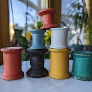 Può includere: Un mucchio di sette bobine di legno dipinte in vari colori, tra cui rosso, blu, giallo, verde e bianco. Le bobine sono disposte a forma di piramide su una superficie bianca.