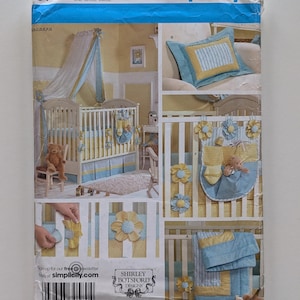 May include: Simplicity sewing pattern for a baby crib set. The pattern features a crib, bedding, and decorative accents in blue and yellow. The pattern includes the text "ONE SIZE" and "SHIRLEY BOTSFORD DESIGNS".