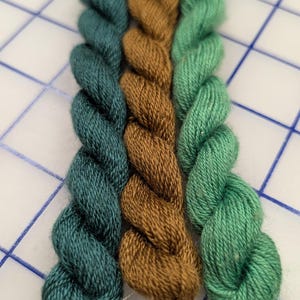May include: Three twisted skeins of embroidery floss arranged vertically. The left skein is a deep teal, the center skein is a warm golden brown, and the right skein is a bright sage green. Each skein features a smooth, multi-ply texture.