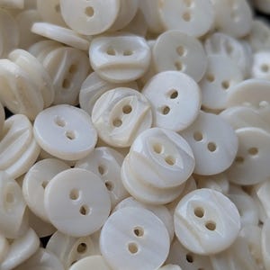 May include: A collection of off-white, round buttons with two holes each. The buttons have a slightly iridescent, pearlescent sheen, and are scattered in a pile. The buttons are likely for sewing or crafting projects.