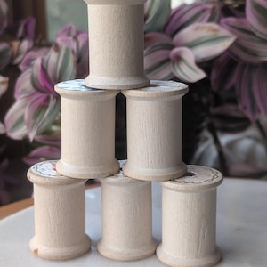 May include: A pyramid of six wooden spools, each with a light beige color and a cylindrical shape. The spools are stacked on a white surface, with a blurred background of green and purple foliage.