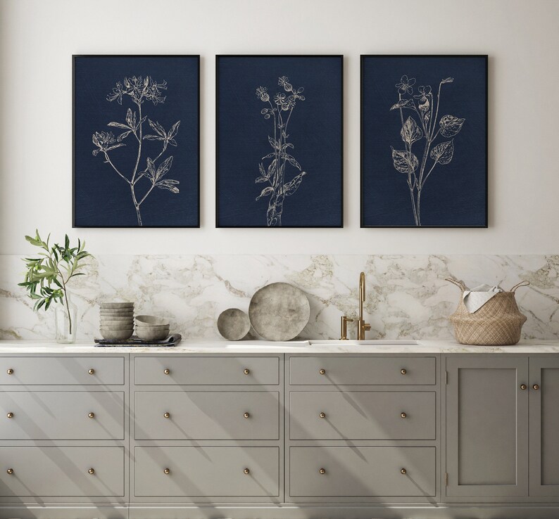 May include: Three framed prints of white line drawings of flowers on a dark blue background. The prints are hanging on a white wall above a gray kitchen cabinet with gold hardware.