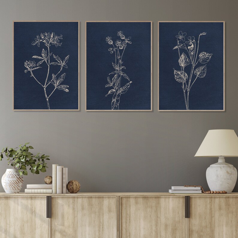 May include: Three navy blue framed prints with white line drawings of botanical illustrations. The prints are hung on a gray wall above a wooden cabinet.