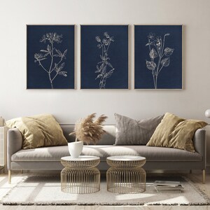 May include: Three framed prints with white line drawings of flowers on a dark blue background. The prints are hanging on a white wall above a gray couch with pillows and a coffee table.