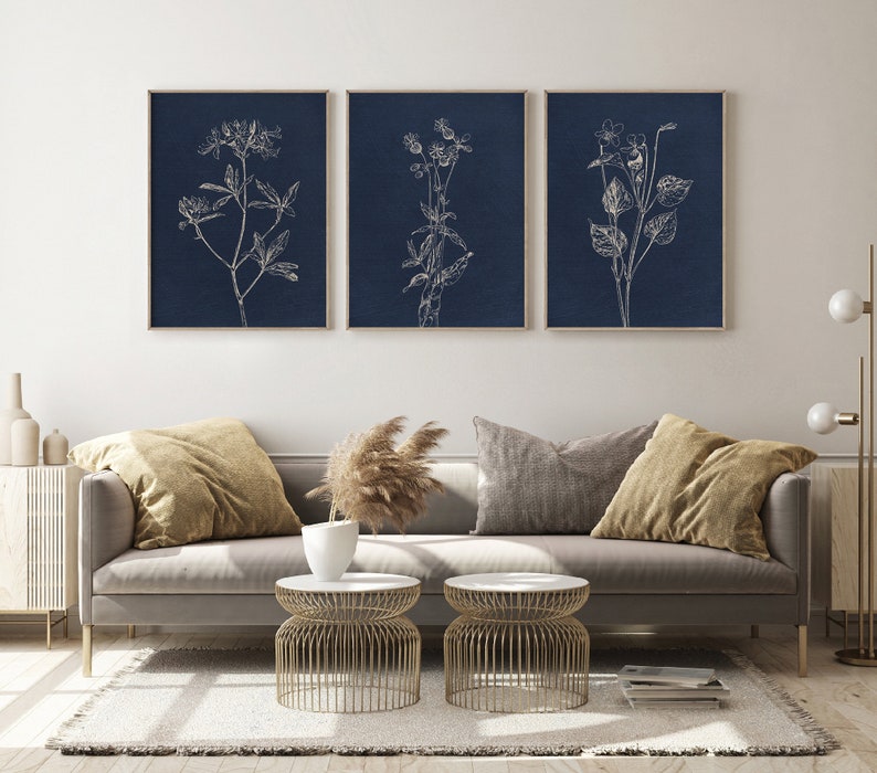 May include: Three framed prints with white line drawings of flowers on a dark blue background. The prints are hanging on a white wall above a gray couch with pillows and a coffee table.