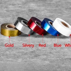 May include: Five rolls of coloured tape in gold, silver, red, blue, and white. Each roll is labelled with its respective colour name. The tape is arranged diagonally on a grey surface.