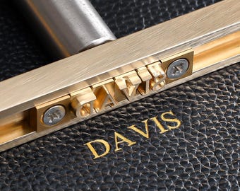 May include: A close-up of a gold-toned metal stamp with the word "DAVIS" engraved on it. The stamp is being used to imprint the name onto a black leather surface.