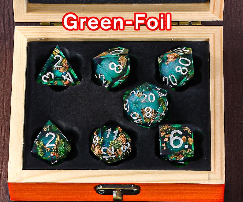 May include: A set of seven green and gold dice with a foil finish. The dice are in a wooden box with a hinged lid. The dice are numbered 1 through 6.
