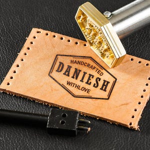 Peut inclure: Une scène d'artisanat du cuir avec une pièce de cuir beige estampillée "HANDCRAFTED DANIESH WITHLOVE" en noir. Un tampon en laiton et un outil noir sont également présents, sur fond sombre.