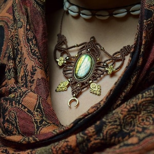 May include: A brown macrame necklace with a large green labradorite gemstone pendant and gold accents. The necklace is worn by a person with a brown patterned scarf.