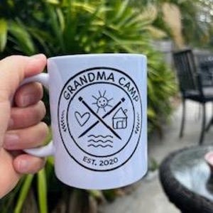 Puede incluir: Taza de cerámica blanca con las palabras "GRANDMA CAMP" en un diseño circular, junto con un corazón, un sol, una casa y agujas de tejer cruzadas. El texto "EST. 2020" está debajo del diseño. La taza tiene un asa blanca.